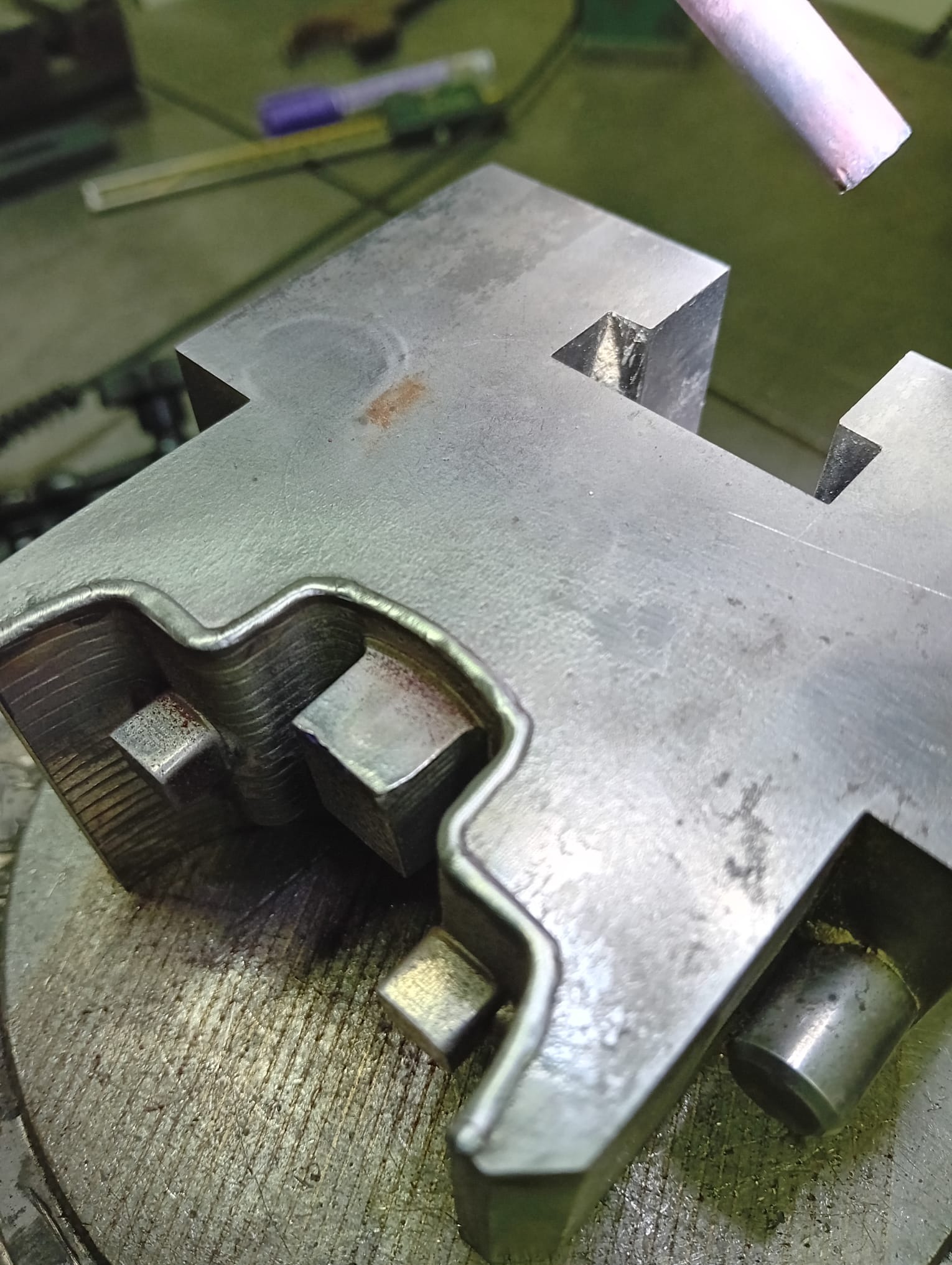 Metalowy element obrabiany, prawdopodobnie uchwyt tokarski, z widocznymi śladami obróbki skrawaniem i spawania, leżący na obrotowym stole roboczym.