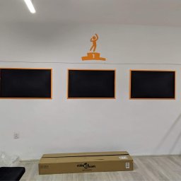 Patryk Józefczyk - Biała ściana biura z trzema czarnymi tablicami w pomarańczowych ramkach i pomarańczową naklejką z motywem osoby na podium, karton na podłodze, częściowo widoczna czarna sofa.