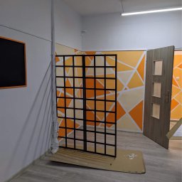 Patryk Józefczyk - Pomarańczowo-białe geometryczne malowanie ścian w biurze, z czarną metalową ścianką działową i otwartymi drzwiami.