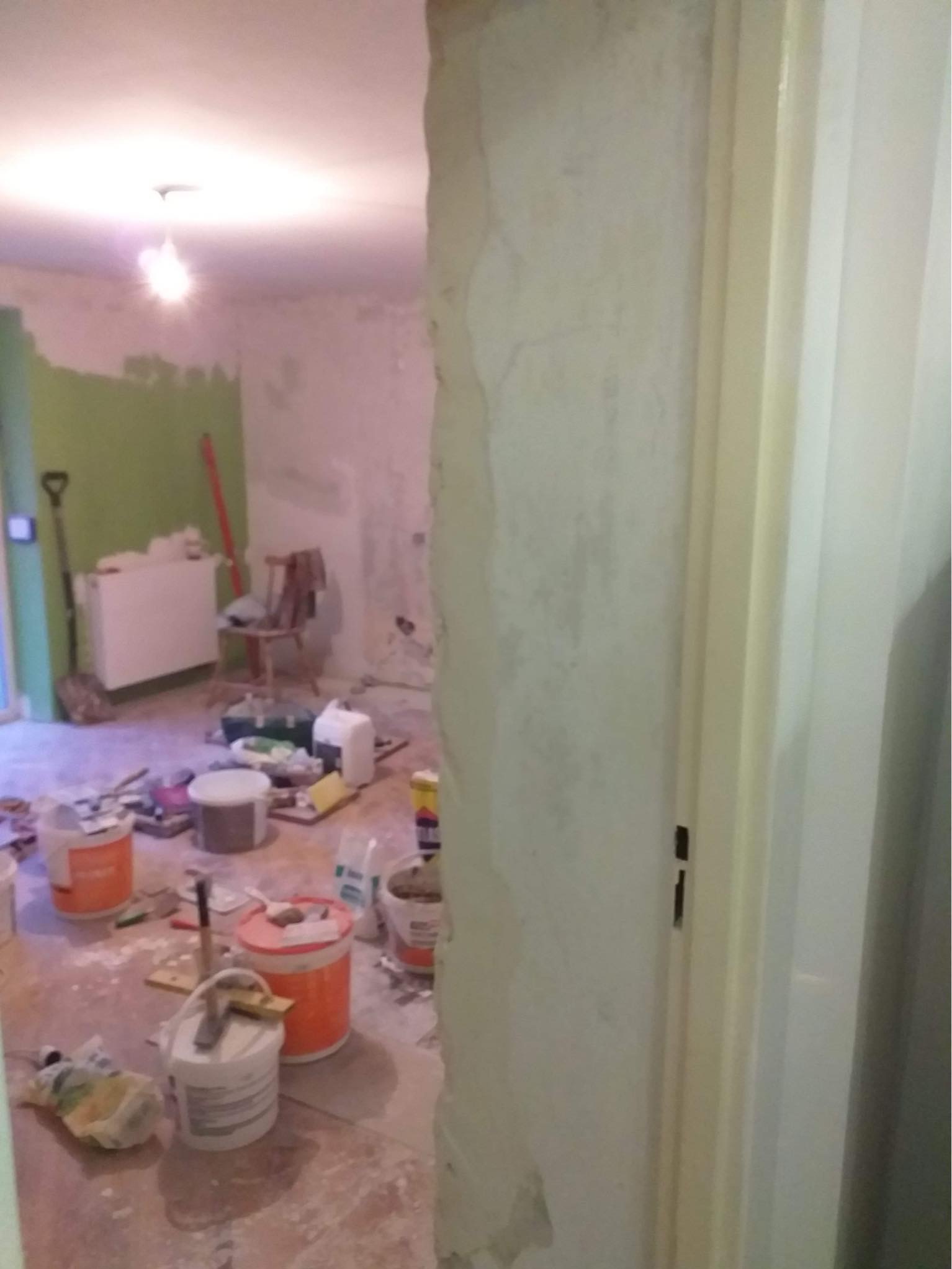 Pokój w trakcie remontu: widoczne wiadra z farbą, narzędzia malarskie, fragment pomalowanej ściany na zielono oraz odsłonięty grzejnik i krzesło w tle, przez niedokończone przejście w ścianie.