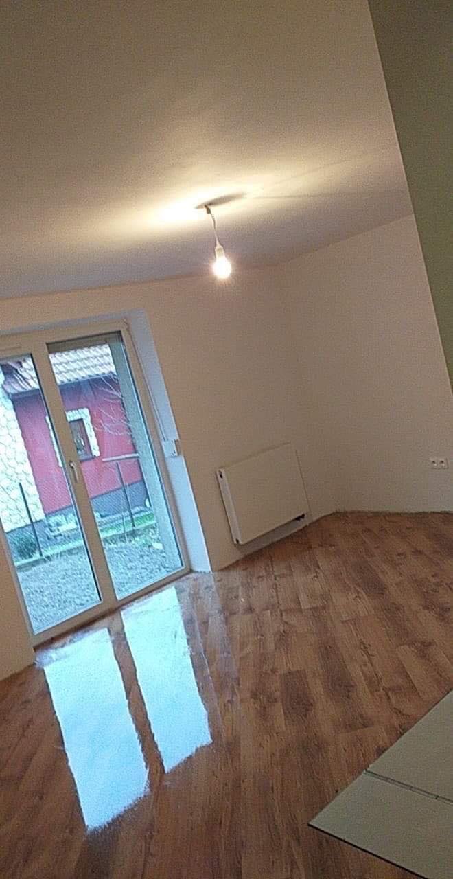 Wnętrze pokoju po remoncie: świeżo pomalowane białe ściany, drewniana podłoga, biały grzejnik pod oknem balkonowym z widokiem na ogród.