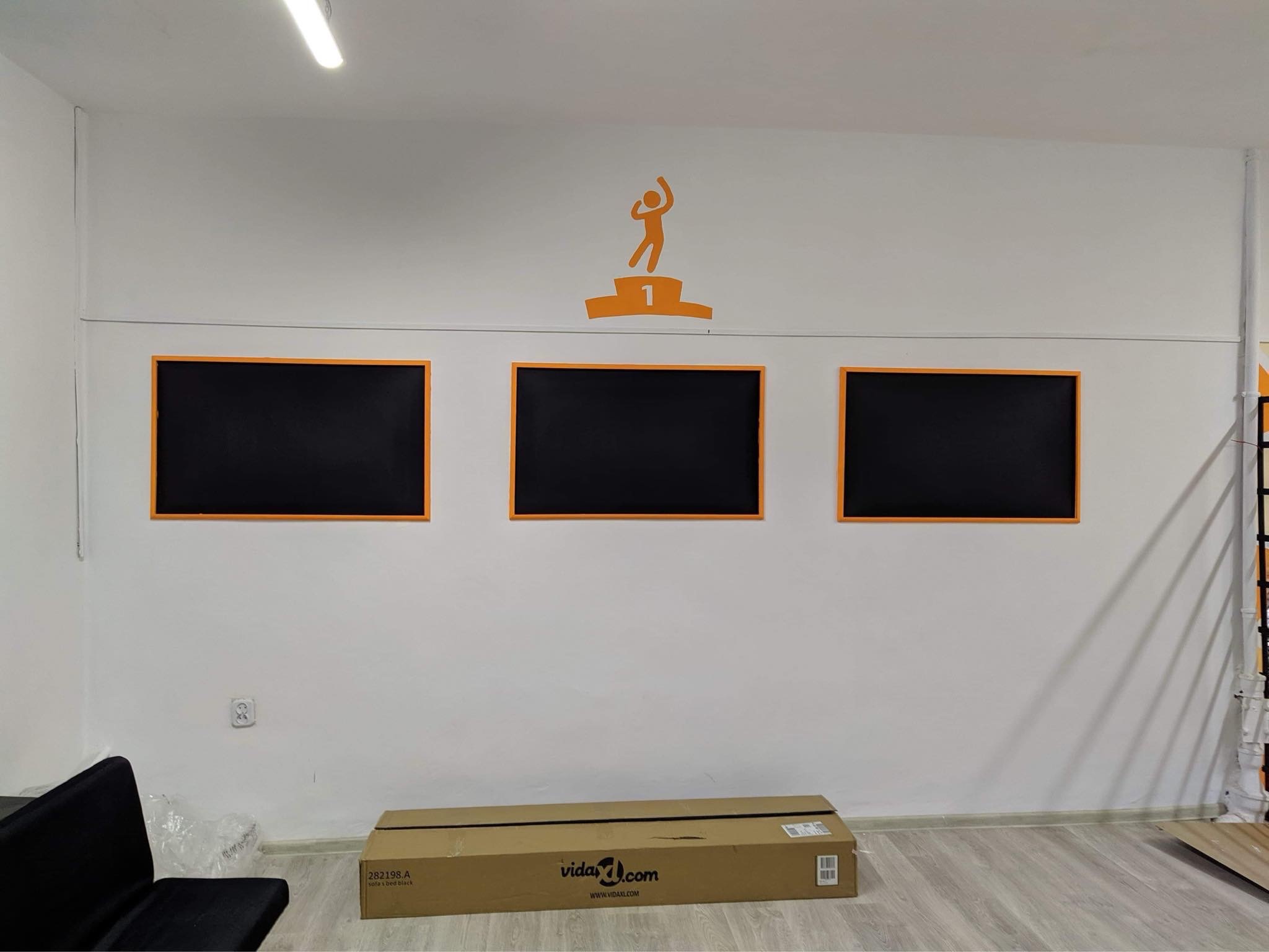 Biała ściana biura z trzema czarnymi tablicami w pomarańczowych ramkach i pomarańczową naklejką z motywem osoby na podium, karton na podłodze, częściowo widoczna czarna sofa.