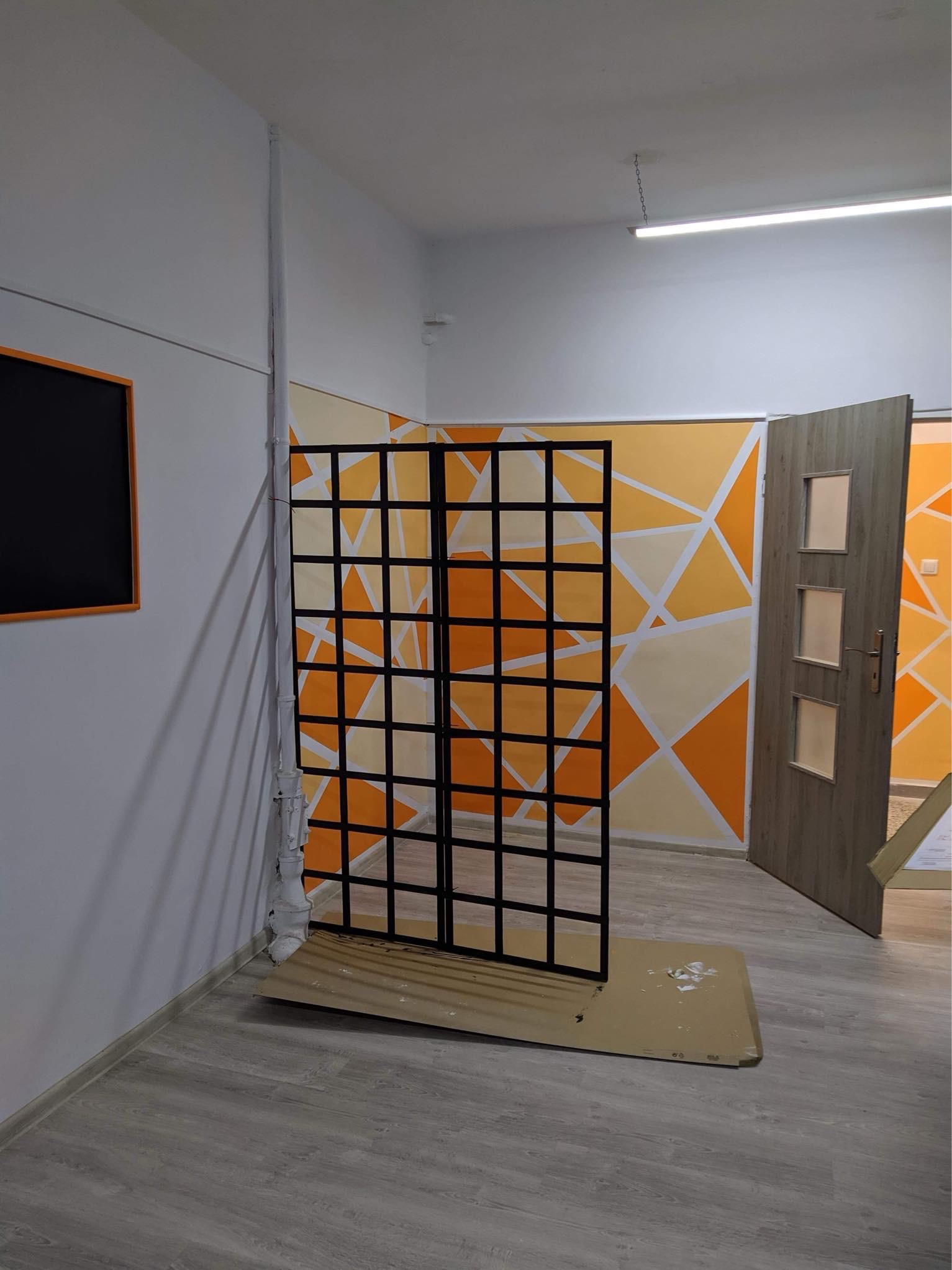 Pomarańczowo-białe geometryczne malowanie ścian w biurze, z czarną metalową ścianką działową i otwartymi drzwiami.