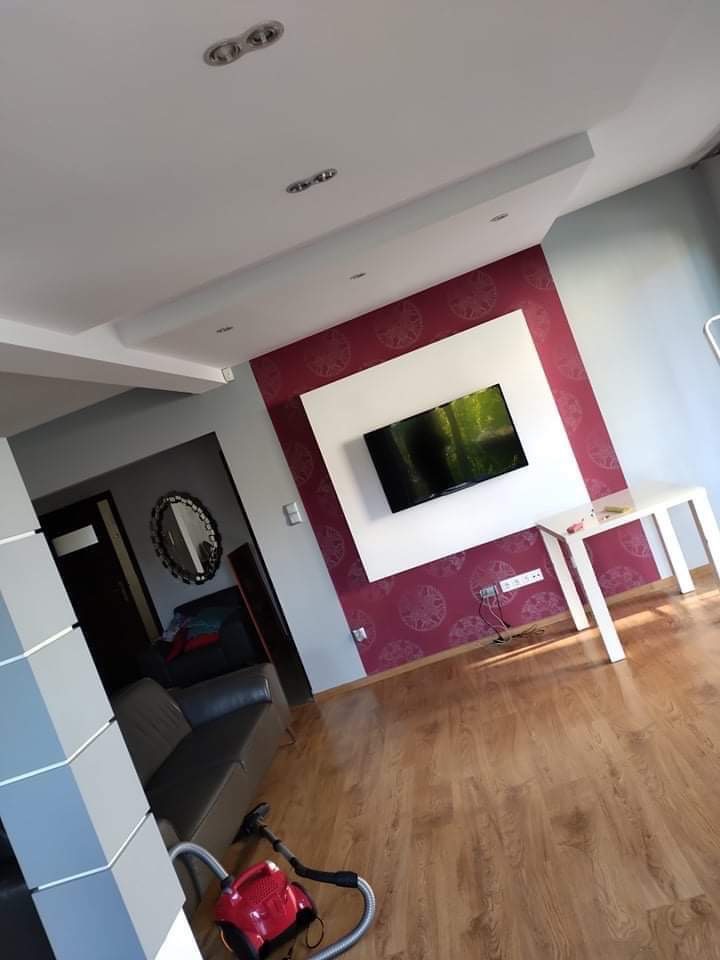 Pokój po remoncie z nowo pomalowaną ścianą w kolorze burgundowym z wzorem, białą ramą wokół telewizora i parkietem, widoczny odkurzacz i fragment kanapy.