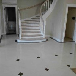 Mrówki polskie przedsiębiorstwo budowlane - Jasna podłoga z marmuru z ciemnymi, kwadratowymi intarsjami oraz białe, drewniane schody kręcone z jasnymi stopniami, widok na otwarte drzwi do pomieszczeń w tle.