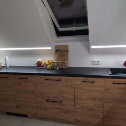 Mrówki polskie przedsiębiorstwo budowlane - Kuchnia w stylu skandynawskim na poddaszu z drewnianymi szafkami, czarnym blatem i oświetleniem LED, widoczne owoce w misce i okno dachowe.