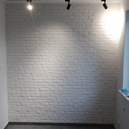 Mrówki polskie przedsiębiorstwo budowlane - Wnętrze po remoncie: ściana z białej cegły, czarne oświetlenie szynowe z reflektorami, szara podłoga z płytek imitujących kamień.