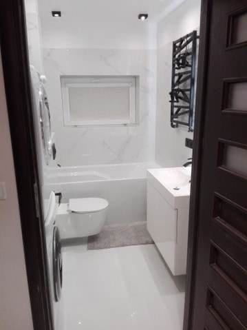 Wnętrze nowoczesnej łazienki z białymi płytkami imitującymi marmur, widok zza uchylonych, ciemnych drzwi. Na ścianie grzejnik drabinkowy w kolorze czarnym, pod oknem wanna, obok podwieszana toaleta...