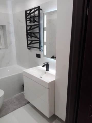 Nowoczesna łazienka z białą umywalką nablatową, czarną armaturą i grzejnikiem drabinkowym w stylu industrialnym, oświetlone lustro z ledami.