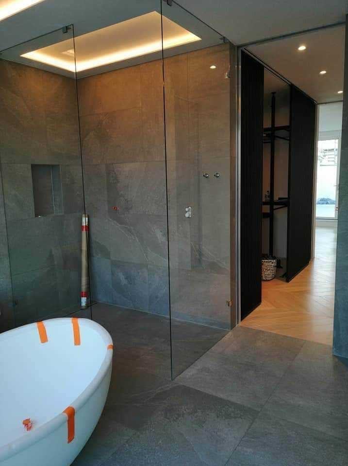 Nowoczesna łazienka z kabiną prysznicową ze szkła, wanną z pomarańczowymi zabezpieczeniami, szarą płytką na ścianach i podłodze oraz wejściem do garderoby z drewnianą podłogą w jodełkę.