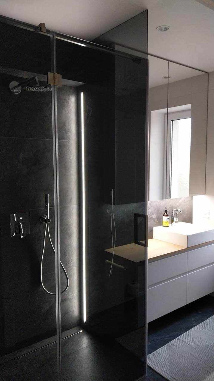 Nowoczesna łazienka z kabiną prysznicową typu walk-in, czarne płytki, pionowe oświetlenie LED w kabinie, biała umywalka nablatowa i szara szafka pod umywalką.
