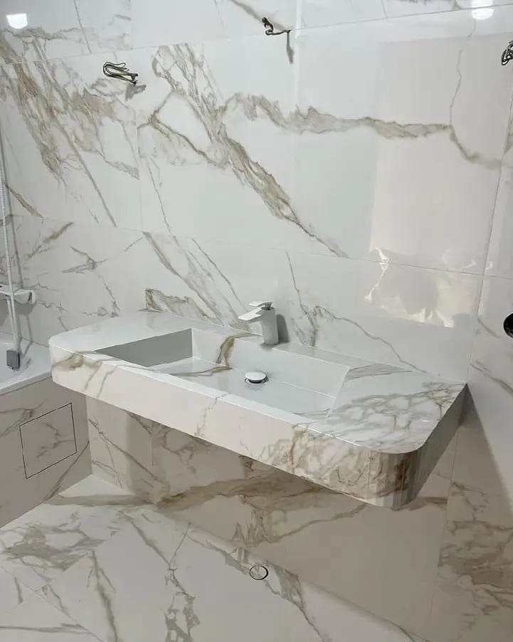 Nowoczesna łazienka z umywalką nablatową z białego marmuru z wyraźnym rysunkiem żył, dopasowanymi płytkami podłogowymi i ściennymi, chromowaną baterią i odpływem.