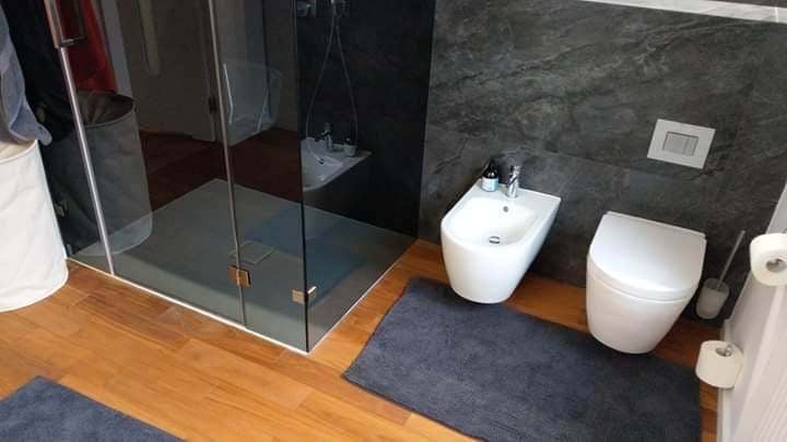 Nowoczesna łazienka z kabiną prysznicową typu walk-in, bidetem i toaletą w kolorze białym, wykończona drewnianą podłogą i szarą, strukturalną ścianą.