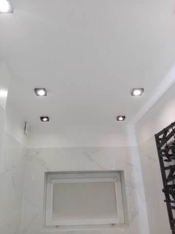 Biały sufit z czterema kwadratowymi lampami LED, białe marmurowe ściany i biała rama okienna z widoczną kratą dekoracyjną po prawej stronie.