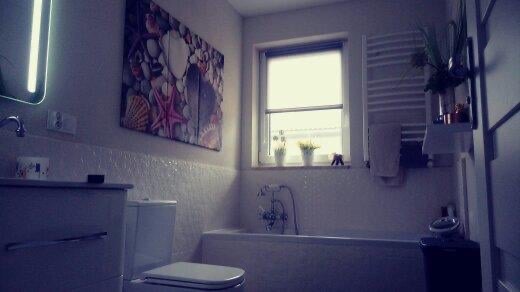 Jasna łazienka z wanną, toaletą, umywalką z szafką, grzejnikiem drabinkowym i oknem z roletą; na ścianie obraz z motywem morskim.