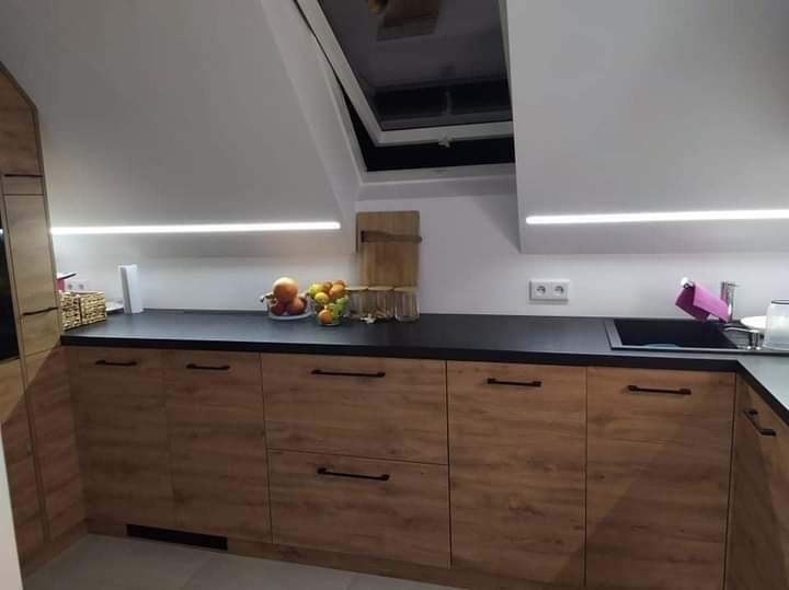 Kuchnia w stylu skandynawskim na poddaszu z drewnianymi szafkami, czarnym blatem i oświetleniem LED, widoczne owoce w misce i okno dachowe.