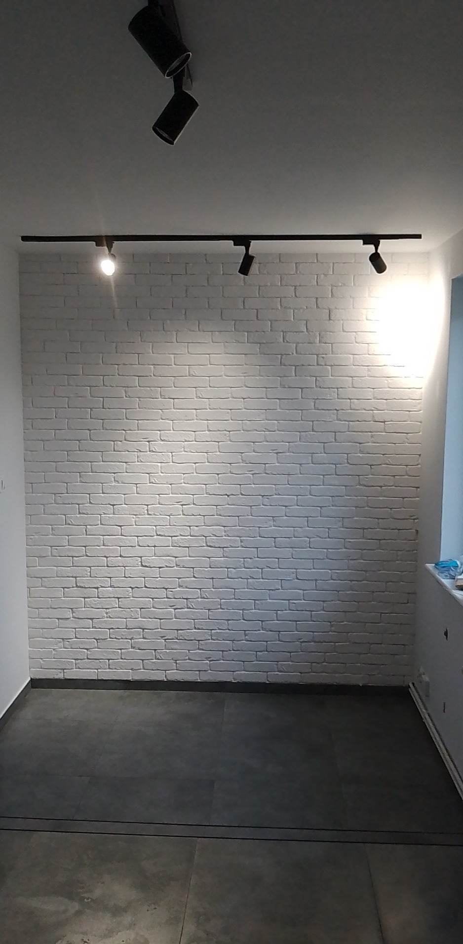 Wnętrze po remoncie: ściana z białej cegły, czarne oświetlenie szynowe z reflektorami, szara podłoga z płytek imitujących kamień.