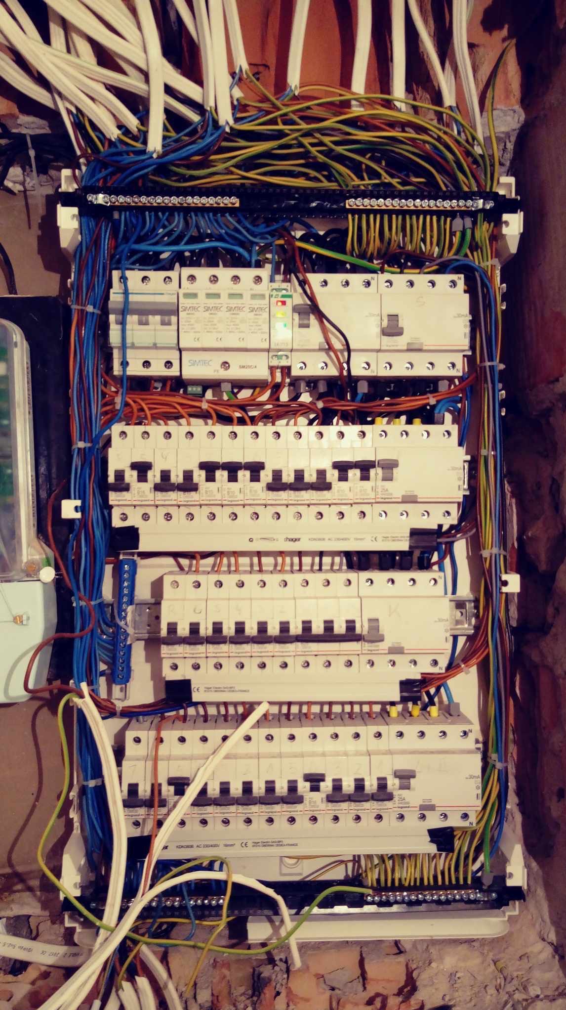 Rozdzielnica elektryczna z licznymi bezpiecznikami i przewodami w różnych kolorach, zamontowana na ścianie z cegły, widoczne elementy marki Simtec i Hager.