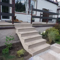 Zewnętrzne schody z płytek prowadzące na taras z drewnianą balustradą, na którym leży pies rasy owczarek niemiecki, obok drabina oparta o ścianę budynku.