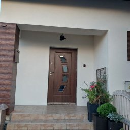 pk-bud - Elewacja domu z brązowymi elementami dekoracyjnymi imitującymi drewno, brązowe drzwi wejściowe z trzema szybkami o nieregularnym kształcie, schody wejściowe wyłożone płytkami, oraz rośliny...