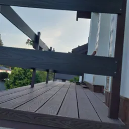 Nowoczesny taras z desek kompozytowych, z widoczną konstrukcją nośną w kolorze ciemnego brązu, oraz z donicą z kwiatami i gołębiem na balustradzie, w tle zabudowa jednorodzinna.