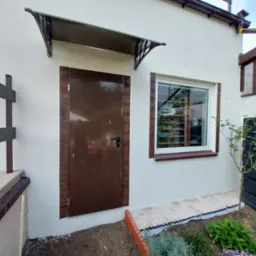 Wejście do budynku z brązowymi drzwiami obramowanymi imitacją drewna, nad drzwiami daszek, obok okno i fragment ogrodu z kamieniami i roślinami.