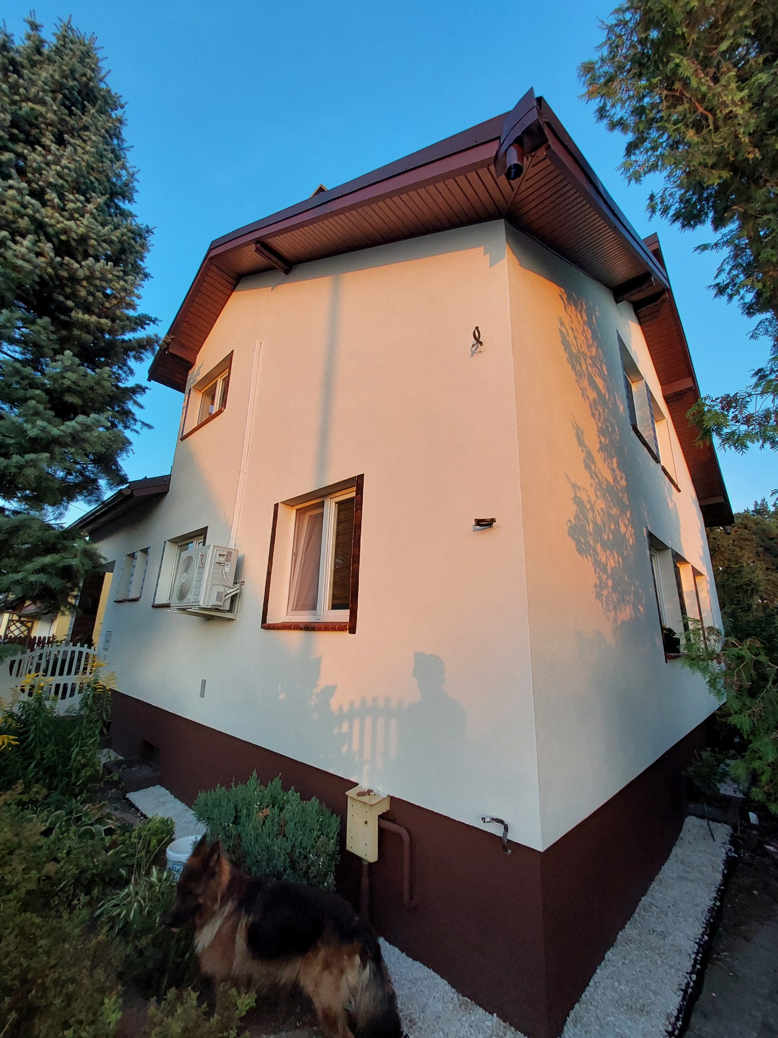 Dwukondygnacyjny dom z nową, kremową elewacją i brązowym cokołem, widoczny klimatyzator i świeżo pomalowane okna, oświetlony ciepłym światłem zachodzącego słońca, z fragmentem ogrodu i psem rasy...