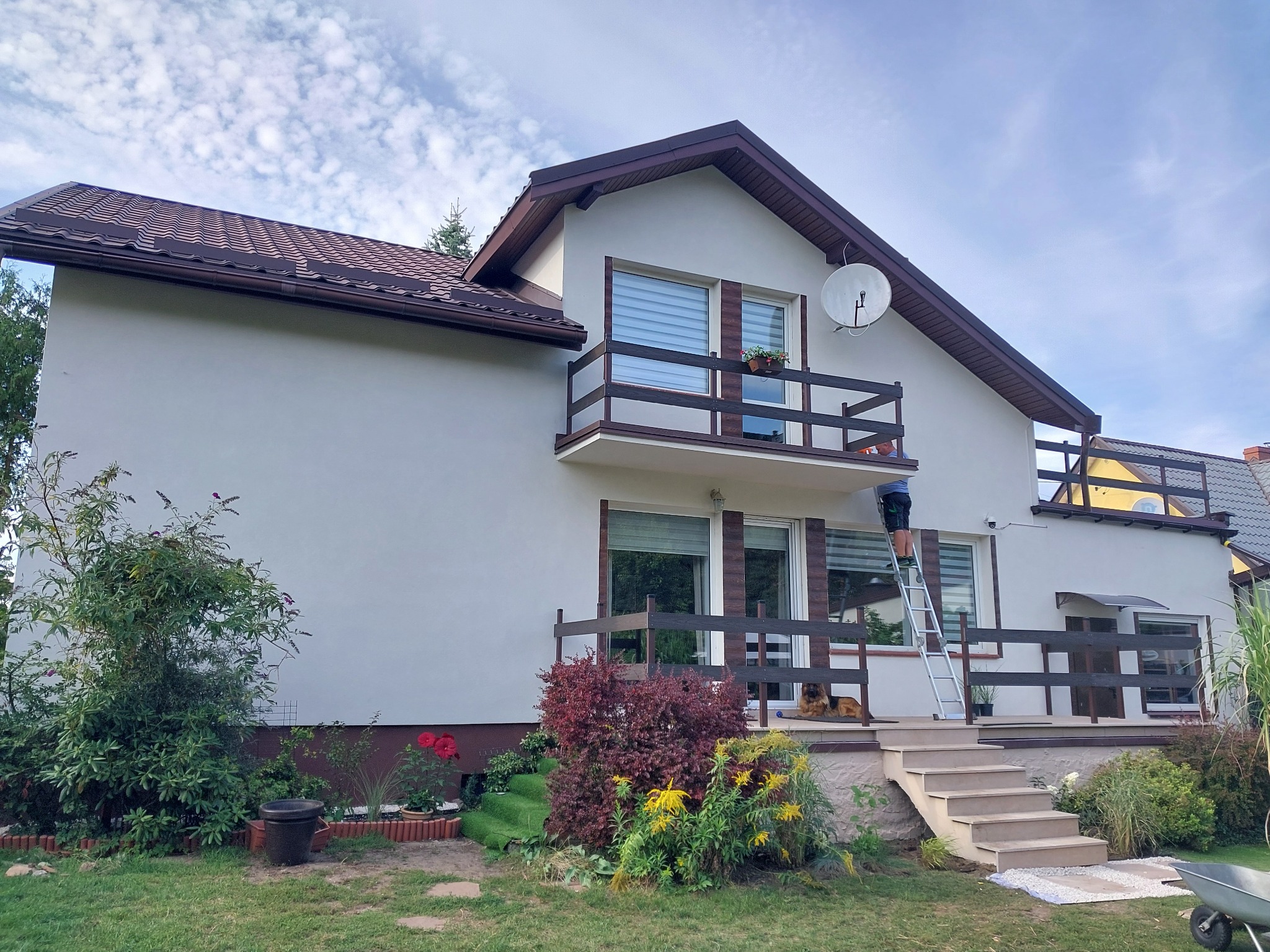 Dwupiętrowy dom z białymi ścianami i brązowym dachem, z balkonem, na którym stoi doniczka z kwiatami; mężczyzna na drabinie maluje elewację; w tle błękitne niebo z chmurami.