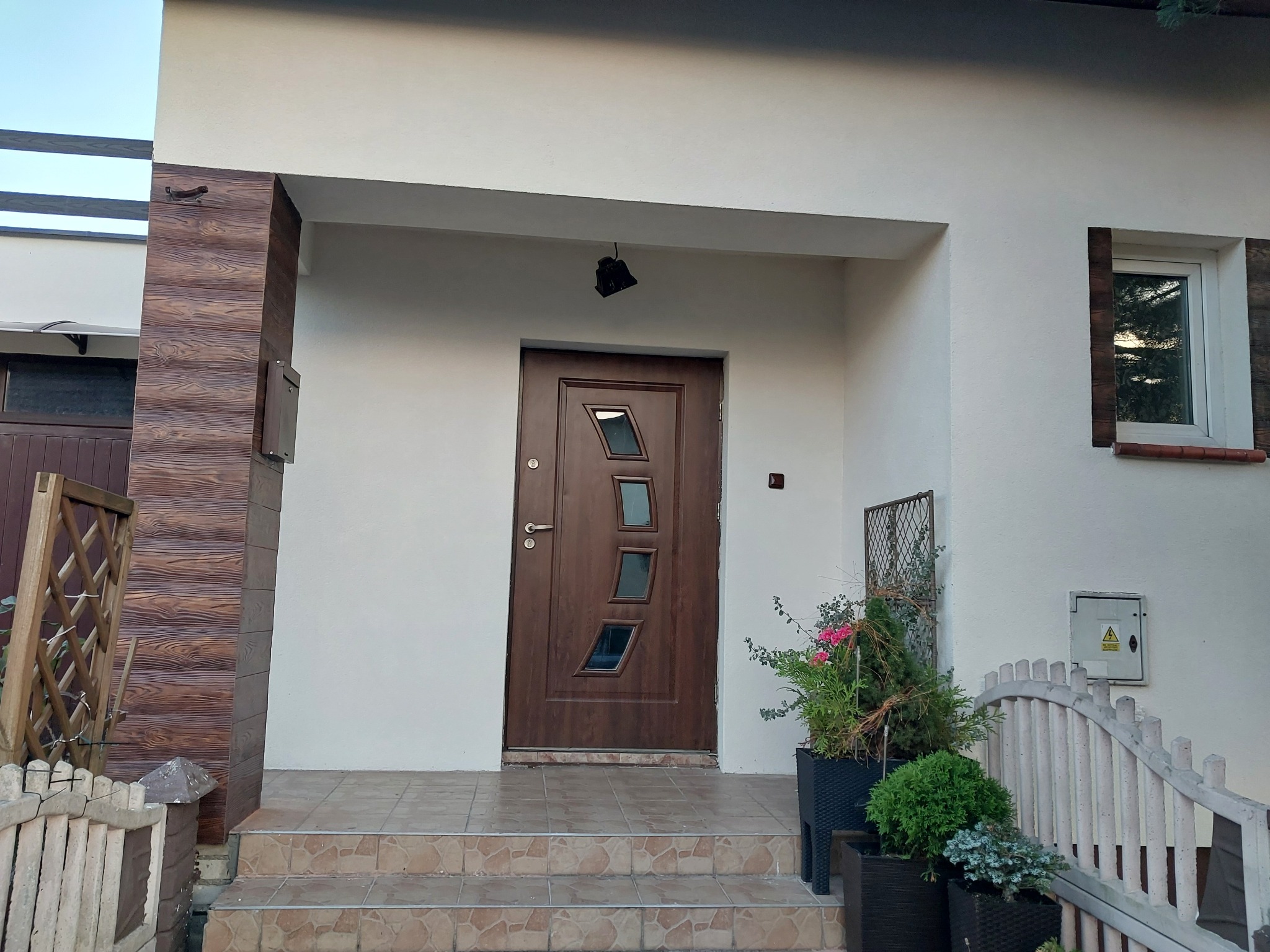 Elewacja domu jednorodzinnego z brązowymi elementami dekoracyjnymi imitującymi drewno, nowoczesne drzwi wejściowe z szybami o nieregularnym kształcie, schody wejściowe wyłożone płytkami i rośliny...