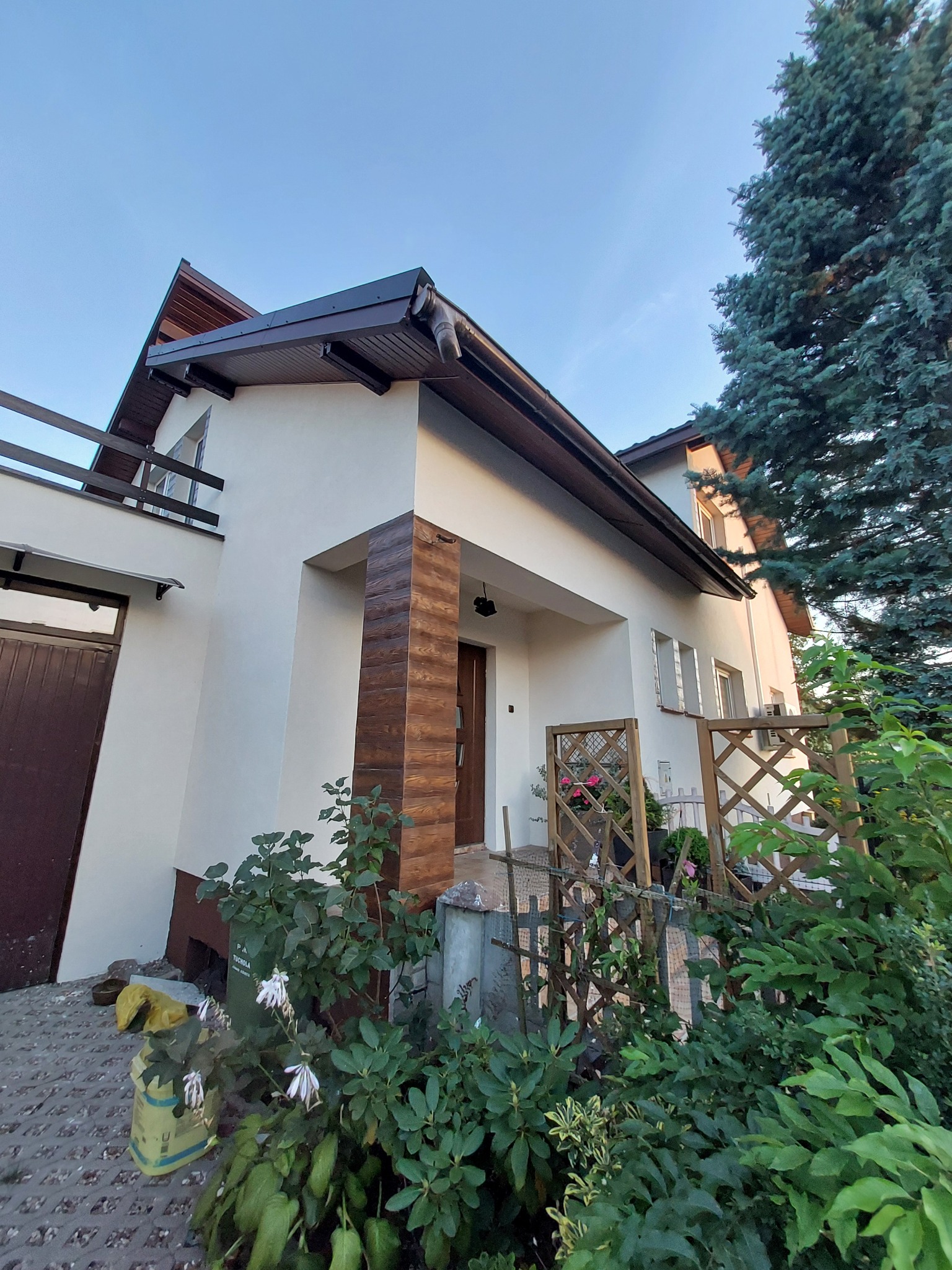 Elewacja domu jednorodzinnego z elementami dekoracyjnymi z drewnopodobnych paneli na filarze wejściowym, świeżo po malowaniu na biało, z widocznym balkonem i zadbaną zielenią w ogrodzie.