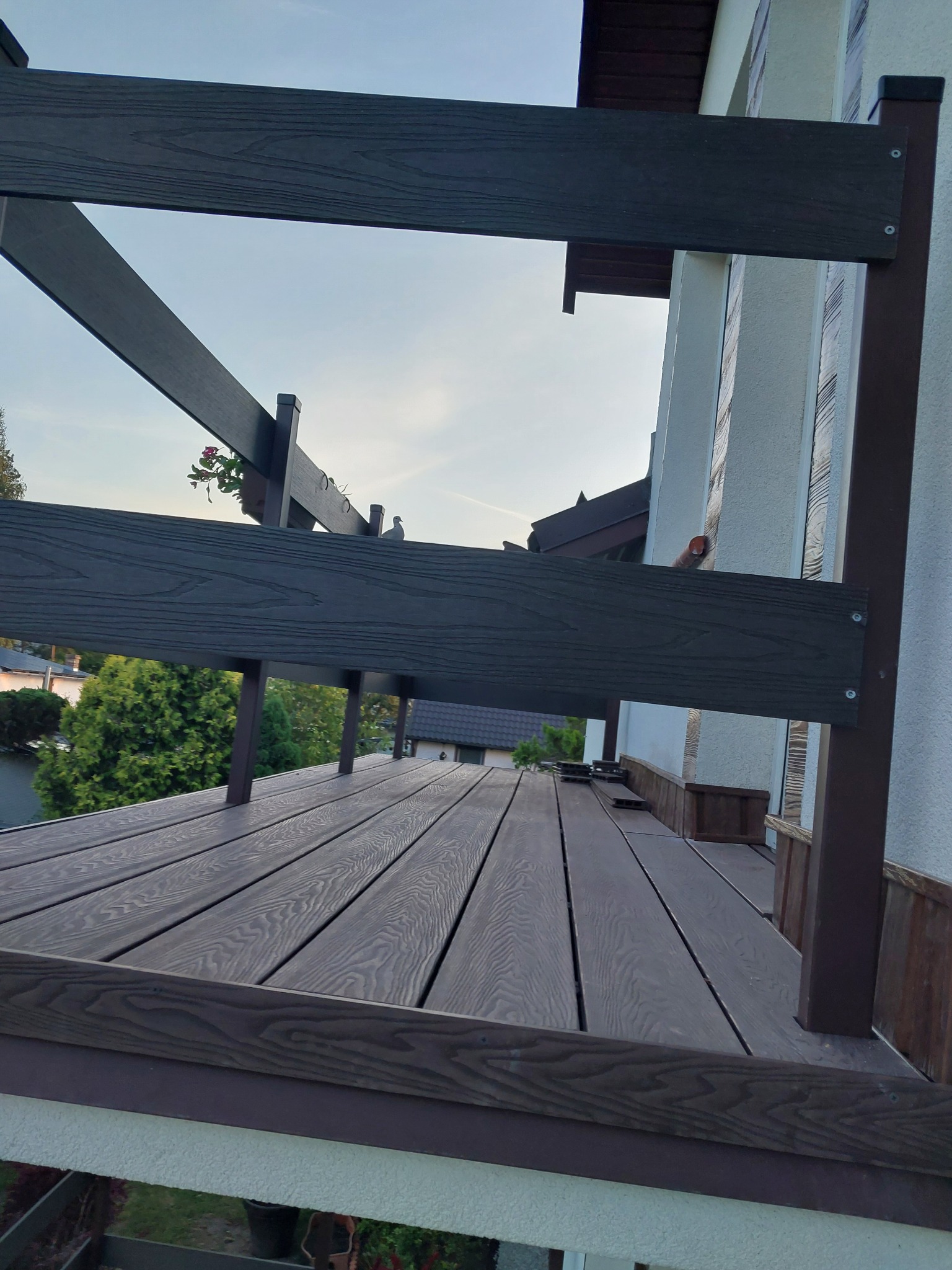 Nowoczesny taras z desek kompozytowych, z widoczną konstrukcją nośną w kolorze ciemnego brązu, oraz z donicą z kwiatami i gołębiem na balustradzie, w tle zabudowa jednorodzinna.