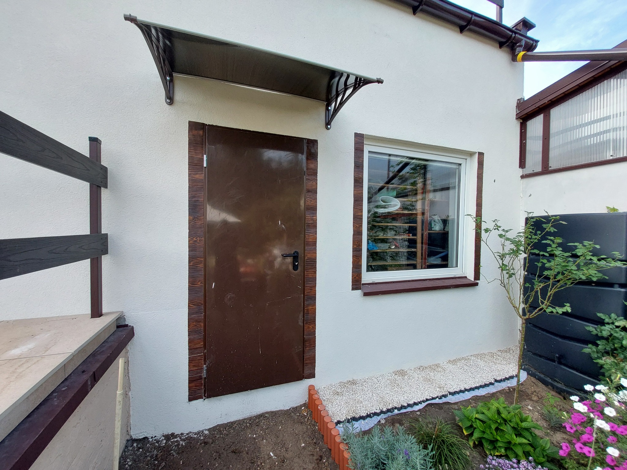 Wejście do budynku z brązowymi drzwiami obramowanymi imitacją drewna, nad drzwiami daszek, obok okno i fragment ogrodu z kamieniami i roślinami.