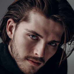 dokuphoto - Portret młodego mężczyzny z ciemnymi włosami zaczesanymi do tyłu, z lekkim zarostem i intensywnym spojrzeniem niebieskich oczu, ubrany w czarny golf na szarym tle.