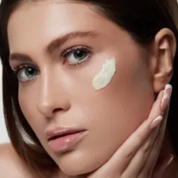 Portret kobiety z kroplą kremu na policzku, delikatnie dotykającej twarzy dłonią z francuskim manicure.