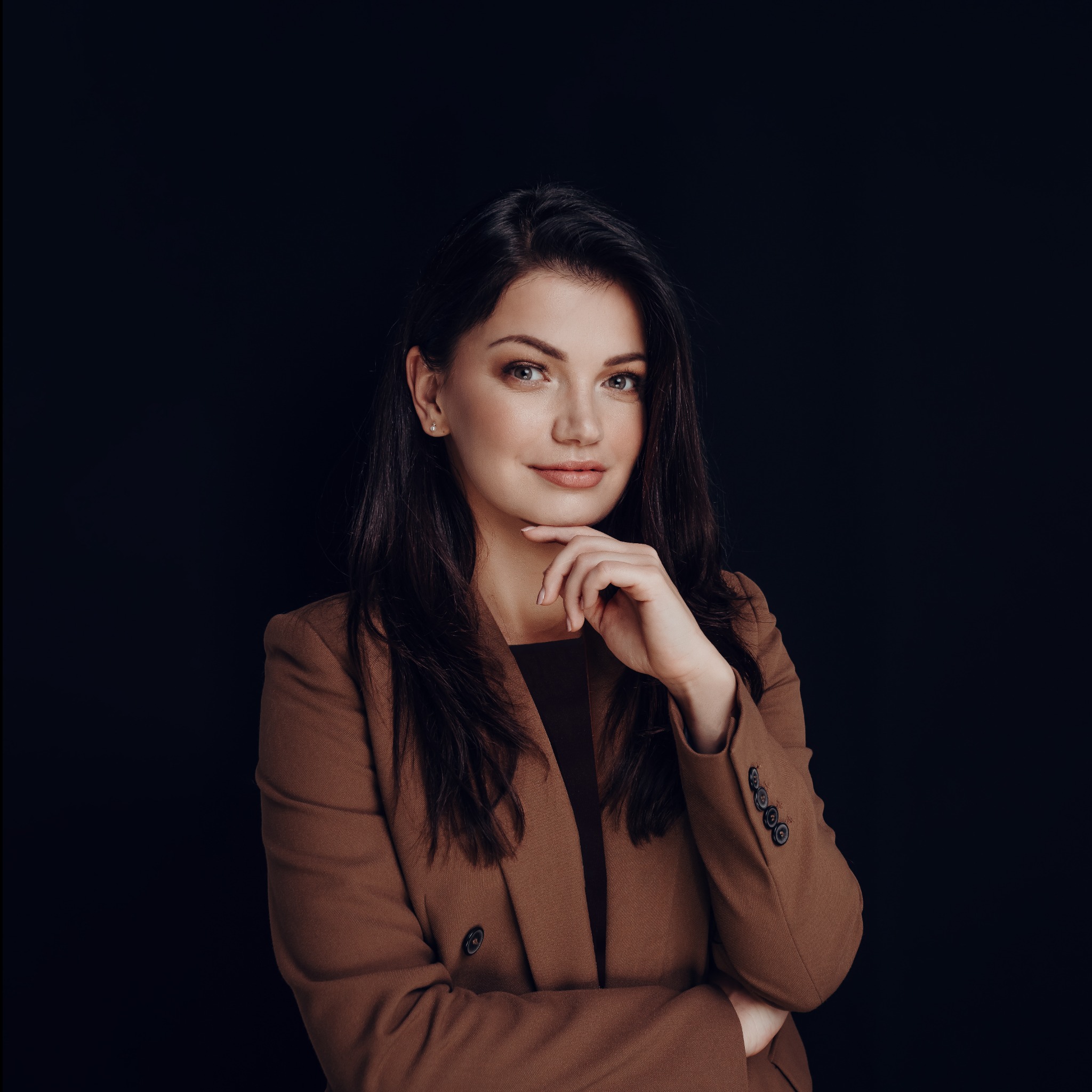 Portret kobiety w brązowym żakiecie na ciemnym tle, z ręką pod brodą i delikatnym uśmiechem.