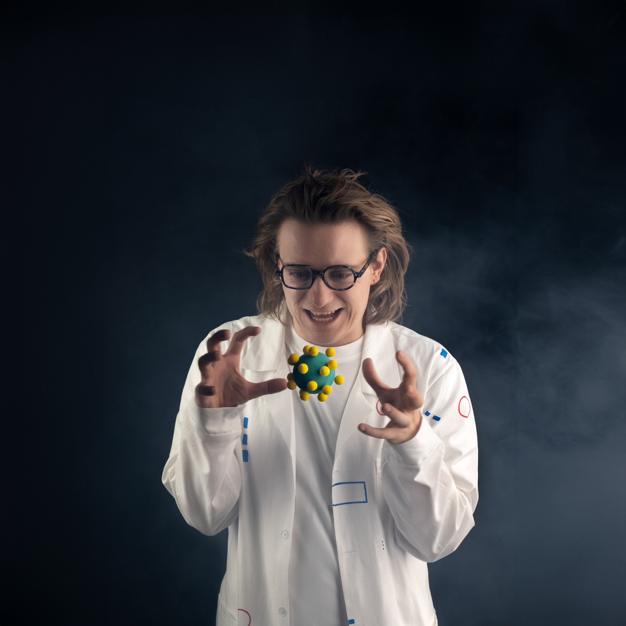 Mężczyzna w okularach i białym fartuchu laboratoryjnym, z entuzjazmem spoglądający na unoszący się w powietrzu model wirusa z zielonej kuli z żółtymi wypustkami, na ciemnym tle.