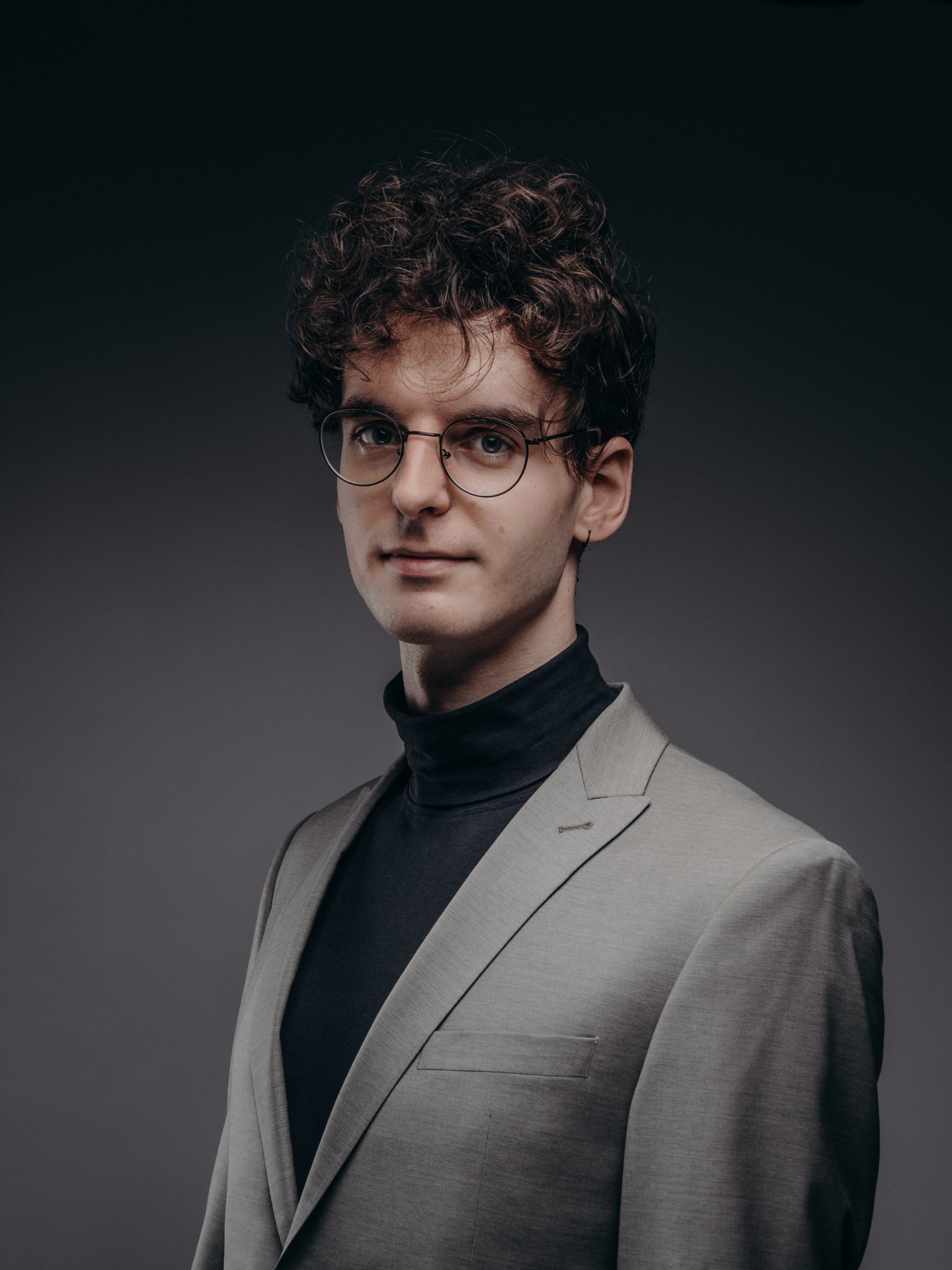 Portret młodego mężczyzny w okularach, czarnym golfie i szarym garniturze na ciemnoszarym tle, z delikatnym uśmiechem i kręconymi włosami.
