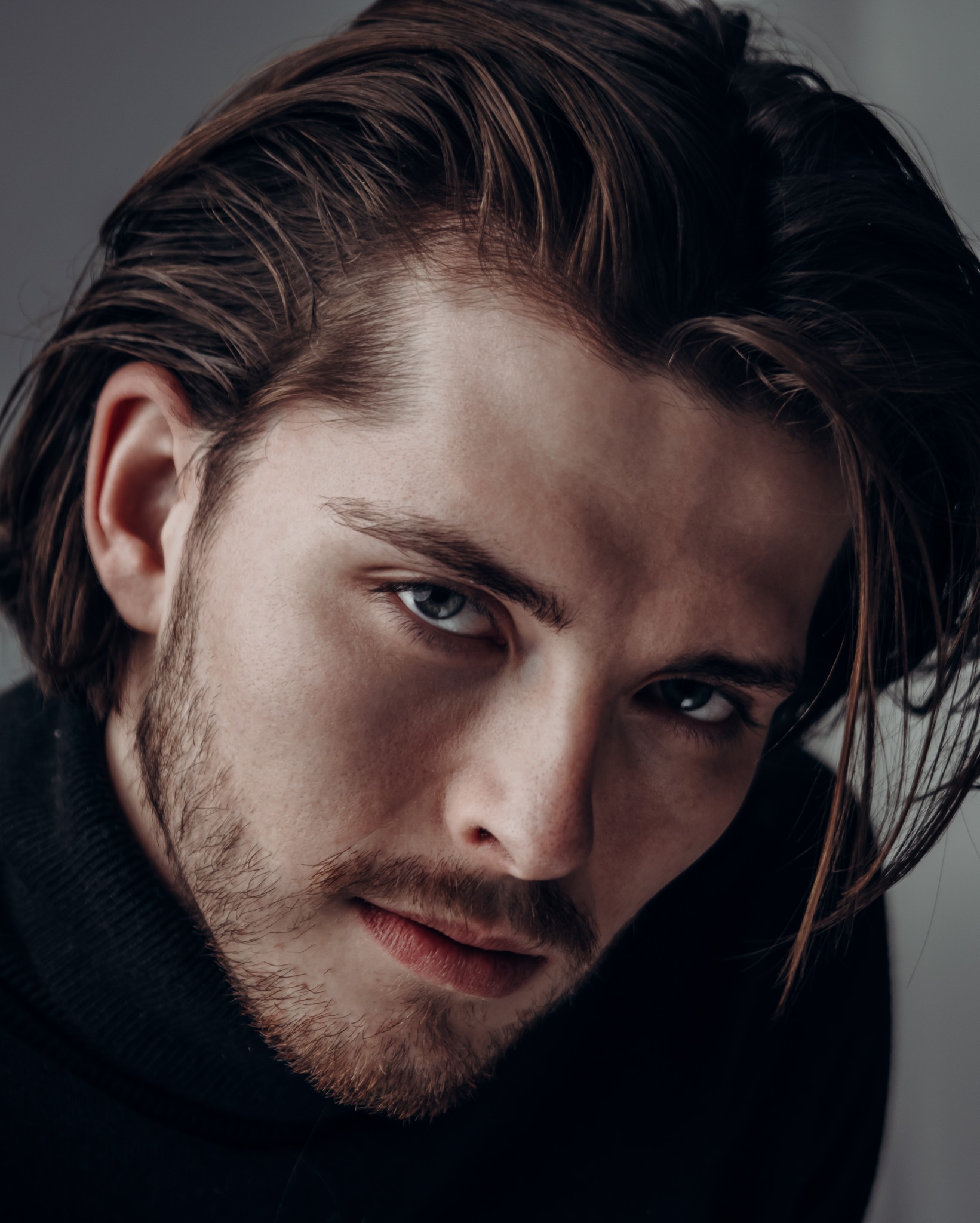 Portret młodego mężczyzny z ciemnymi włosami zaczesanymi do tyłu, z lekkim zarostem i intensywnym spojrzeniem niebieskich oczu, ubrany w czarny golf na szarym tle.