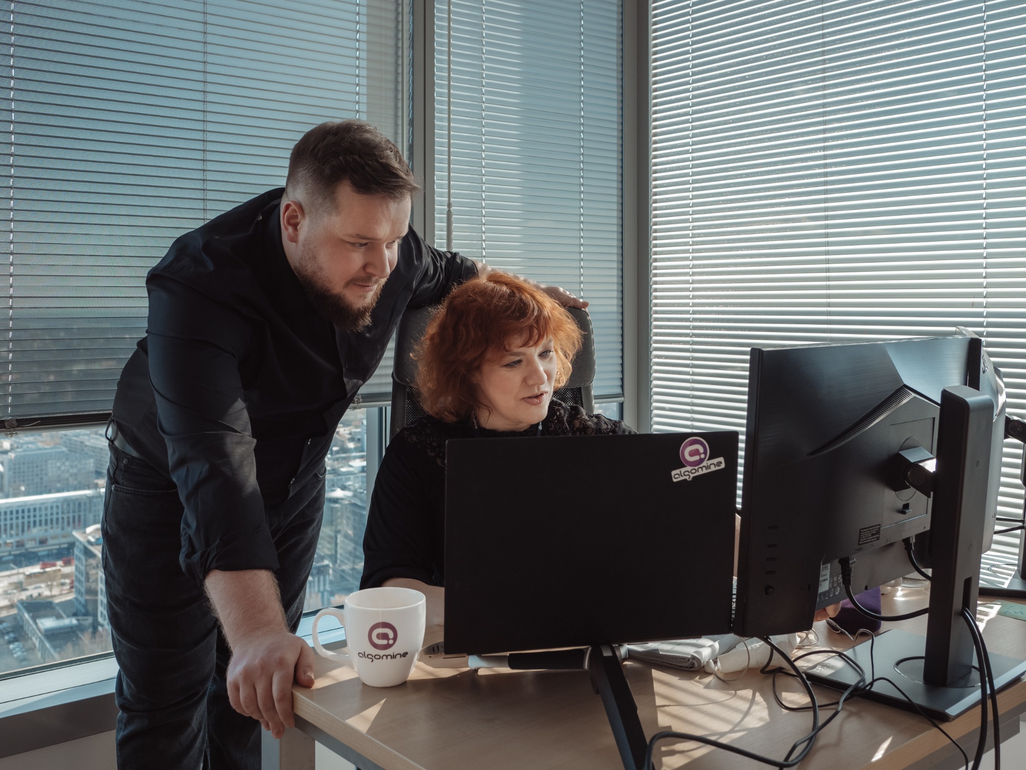 Mężczyzna w czarnej koszuli spogląda na kobietę z rudymi włosami pracującą na laptopie w biurze z żaluzjami, na biurku kubek z logo firmy.