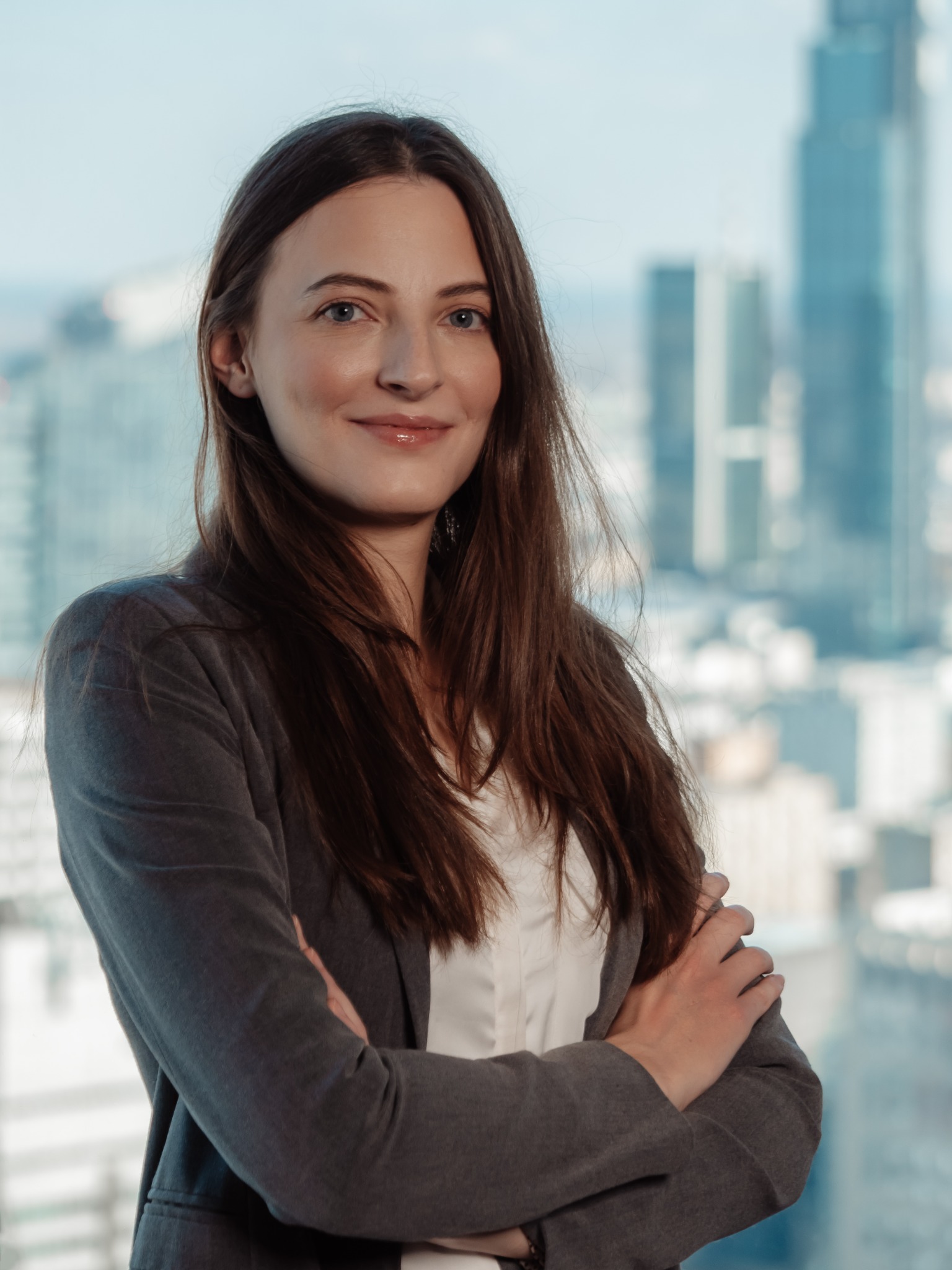 Portret uśmiechniętej kobiety w szarej marynarce, z rękami skrzyżowanymi na piersiach, na tle rozmytych wieżowców widzianych przez okno.