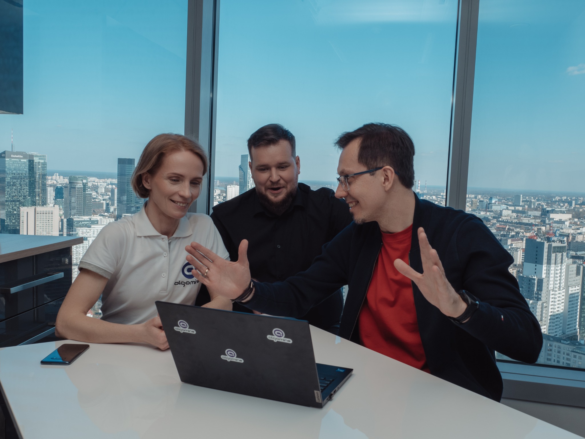 Trzy osoby w biurze z widokiem na panoramę miasta, rozmawiające i gestykulujące nad laptopem z naklejkami logotypu. Kobieta w polo z logo firmy, mężczyźni w koszuli i marynarce.