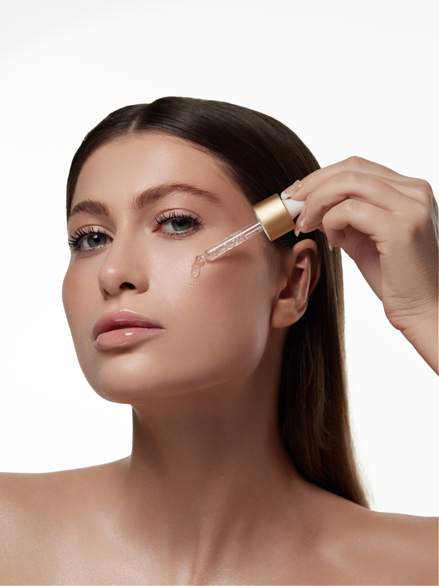 Portret kobiety aplikującej serum z pipety na policzek, na białym tle.