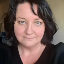 Portret kobiety o ciemnych, kręconych włosach i zielonych oczach, ubranej w czarną bluzkę z bufkami na ramionach, na rozmytym tle.