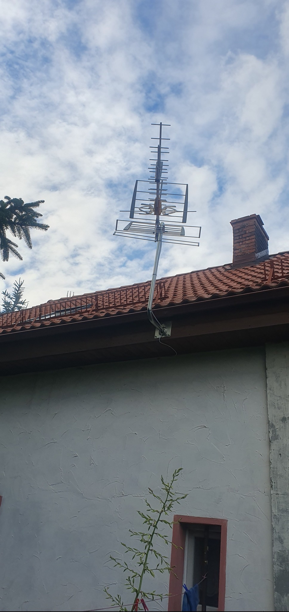 Zewnętrzna antena telewizyjna zamontowana na dachu budynku z czerwoną dachówką, na tle błękitnego nieba z chmurami. Widoczny komin i fragment ściany budynku.