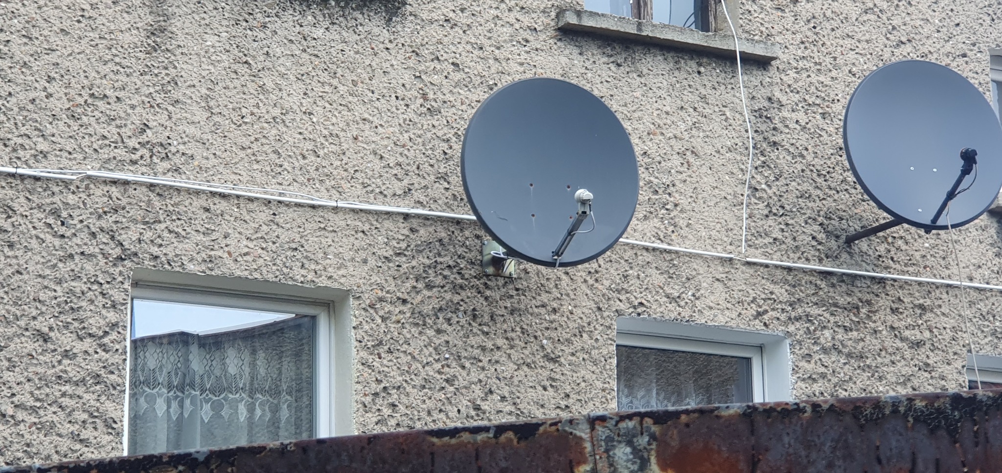 Dwie szare anteny satelitarne zamontowane na szorstkiej, jasnoszarej elewacji budynku, z widocznymi białymi kablami i oknami.