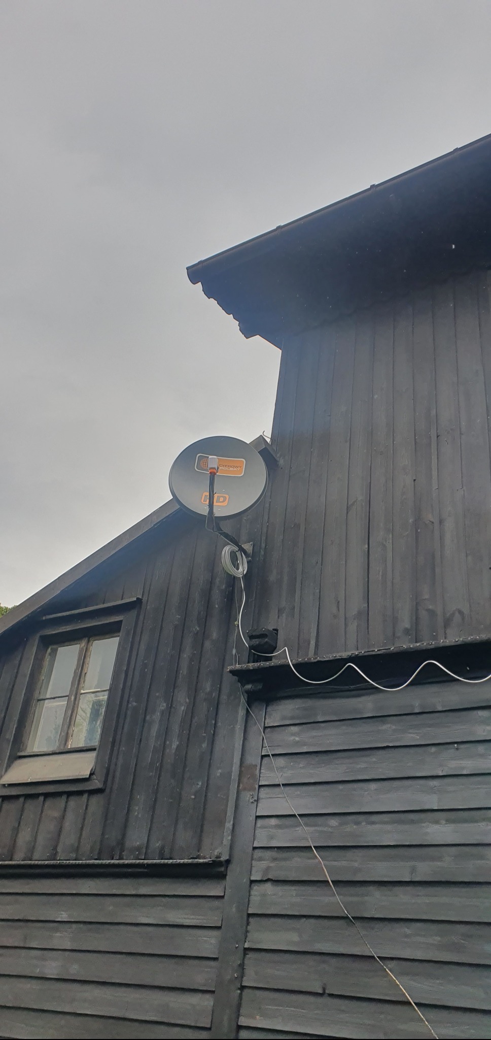 Antena satelitarna HD zamontowana na ciemnej, drewnianej ścianie budynku. Widoczne okno i kabel antenowy. Pochmurne niebo w tle. Kąt ujęcia z dołu.