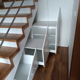 Maciej Kukuła - Białe szafki i szuflady w zabudowie pod schodami z drewnianymi stopniami i metalową barierką, widoczne otwarte półki i wysunięta szuflada.