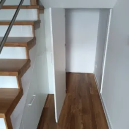 Schody drewniane z zabudową szaf wnękowych pod nimi, białe fronty szafek, otwarte drzwi do przestrzeni schowka, parkiet drewniany.