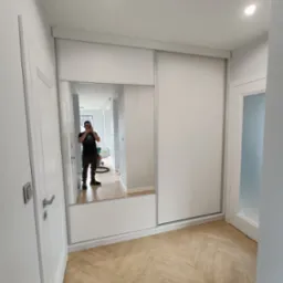 Biała szafa przesuwna z lustrem w przedpokoju, widoczny parkiet w jodełkę i odbicie fotografa.
