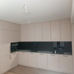 Maciej Kukuła - Jasna, nowoczesna kuchnia z beżowymi szafkami, czarnym blatem i panelem ściennym, widoczne niedokończone podłączenia elektryczne na suficie.